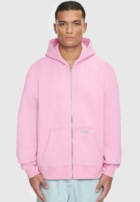Light pink zip-up hoodie made of soft fabric. Features a spacious hood, front pockets, and ribbed cuffs. Minimalistic design with no patterns.