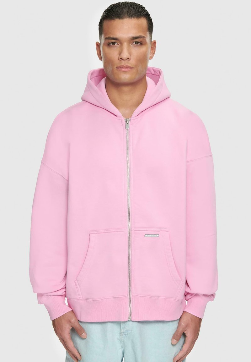 Light pink zip-up hoodie made of soft fabric. Features a spacious hood, front pockets, and ribbed cuffs. Minimalistic design with no patterns.