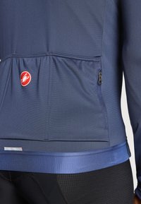 Haut à manches longues navy avec une surface texturée, taille élastique, logo rouge et une poche zippée sur le côté.