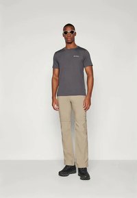 Άνδρας ντυμένος με σκούρο γκρι μπλουζάκι Columbia, μπεζ παντελόνια convertible, μαύρα παπούτσια και λευκά γυαλιά ηλίου, που στέκεται μπροστά σε ουδέτερο φόντο.