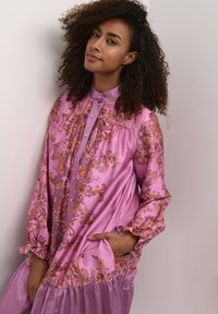 Ružová blúzka s vzormi, ktorá má volánové detaily, vysoký golier a dlhé rukávy s elastickými manžetami. Mäkkučký materiál so kvetinovými akcentmi.