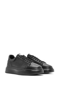 Een paar zwarte leren sneakers met dikke zolen en zwarte veters, naast elkaar geplaatst op een witte achtergrond.