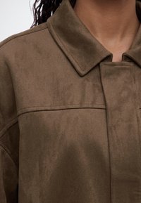 Gros plan d'une personne portant une veste en daim marron avec un col large et des détails de couture visibles.