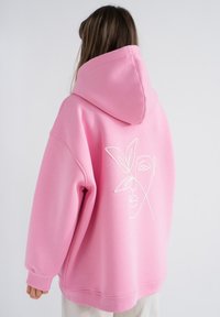 Felpa oversized rosa realizzata in un materiale morbido, con un disegno di volto e foglia tracciato in bianco sul retro. Include un cappuccio ampio.
