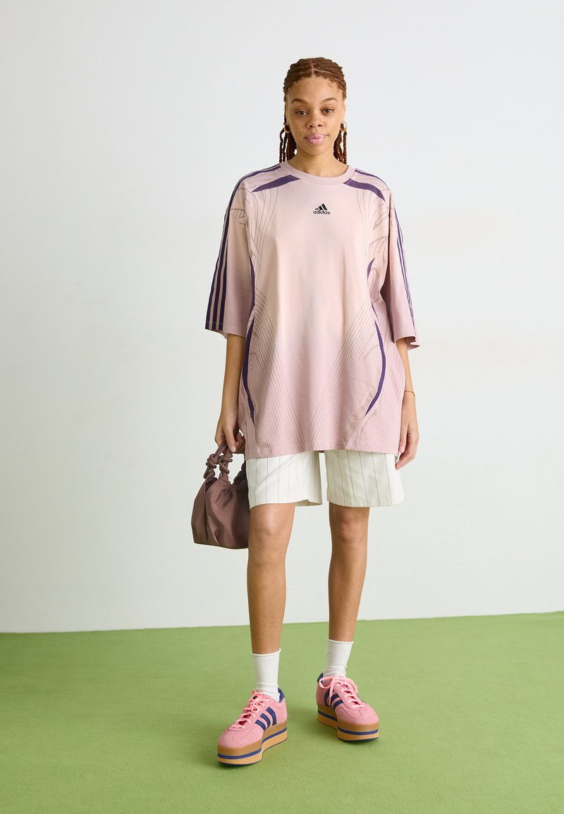 Przesadnie duża różowa koszulka sportowa z fioletowymi akcentami, białe spodenki w paski, różowe trampki na platformie i mała brązowa torebka.