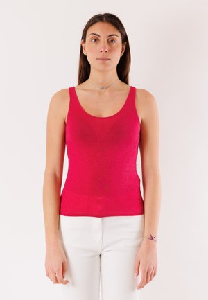 Donna con canotta rosa acceso senza maniche e pantaloni bianchi, in piedi davanti a uno sfondo chiaro uniforme, rivolta verso l'obiettivo.