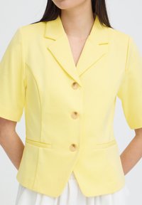 Femme portant un blazer jaune à manches courtes avec col à revers, trois boutons et deux poches à fente sur le devant.