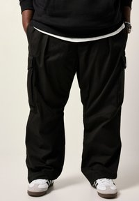 Schwarze Cargo-Hosen mit lockerem Schnitt, ausgestattet mit seitlichen Taschen und einer glatten Textur. Kombiniert mit weißen und grauen Sportschuhen.