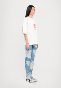 Hvit oversized t-skjorte med grafisk trykk, blå bukser med sky-mønster og sølvfargede heeled mules. Glatte teksturer og avslappet design.
