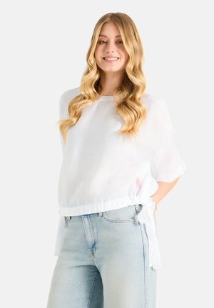 Junge Frau mit langen blonden welligen Haaren, die eine lockere weiße Bluse trägt, die seitlich gebunden ist, und hellblaue Jeans, lächelnd vor einem schlichten Hintergrund