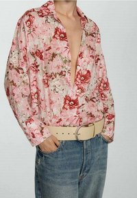 Camicia floreale rosa e bianca, caratterizzata da una vestibilità ampia, colletto e maniche lunghe, abbinata a jeans a vita alta e una cintura beige.