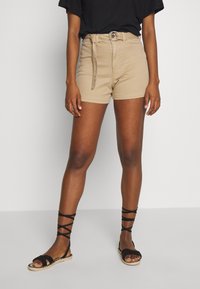 Person wearing beige belted shorts and black lace-up sandals, standing against a plain light gray background.