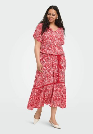 Femme portant une robe rouge à motifs, manches courtes, avec une ceinture nouée à la taille, debout et regardant vers le bas, chaussée de talons blancs à bout pointu.