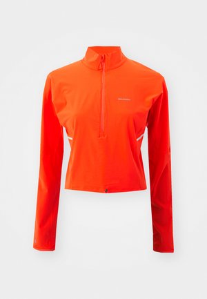 Jasnopomarańczowa sportowa bluzka z długim rękawem i ćwierć zamkiem, wykonana z gładkiego, elastycznego materiału. Posiada małe logo oraz elementy odblaskowe.