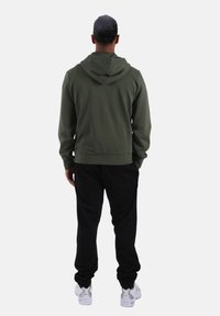 Felpa con cappuccio verde scuro realizzata in tessuto morbido, dotata di cappuccio spazioso e polsini a costine, abbinata a pantaloni neri e sneakers bianche.