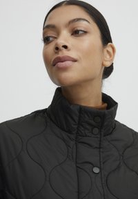Chaqueta negra acolchada con cuello alto, que presenta patrones de costura circulares y botones a presión negros, usada sobre una blusa marrón.