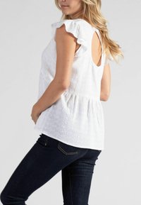 Weißes Baumwolloberteil mit Rüschensäumen, gerafftem Taillenbereich und einem Ausschnitt am Rücken. Das Lochstickmuster sorgt für Struktur und Details. Kombiniert mit dunklen Jeans.