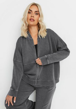 Harmaa vetoketjullinen huppari rentoa istuvuutta, jossa on ribbetty kaulus ja hihansuut, ommeltuja yksityiskohtia ja pehmeä tekstuuri. Yhdistetty samansävyisiin housuihin.