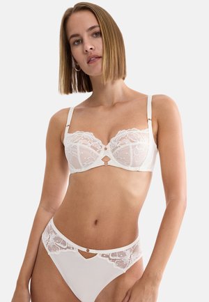 Woman with shoulder-length hair wearing white lace lingerie set with sheer floral details, standing against a plain light background.