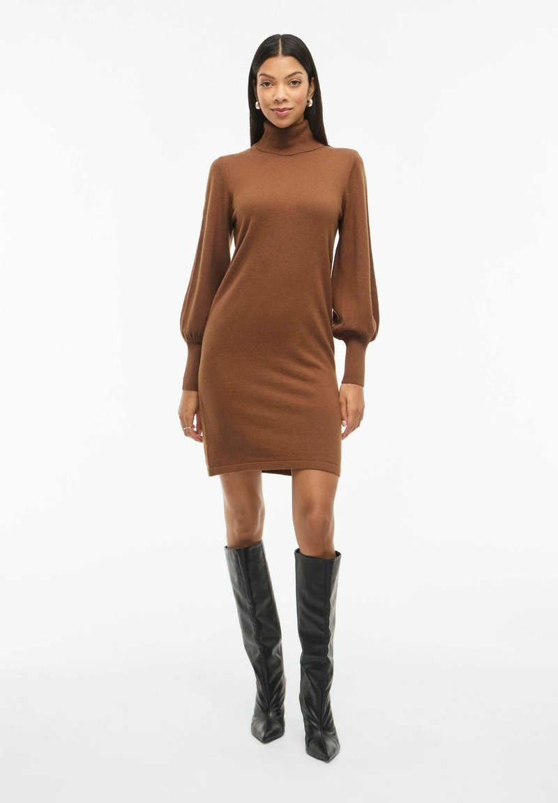 Donna che indossa un vestito lungo a maniche effetto turtleneck marrone con maniche a sbuffo e stivali neri al ginocchio, in piedi su uno sfondo bianco semplice.