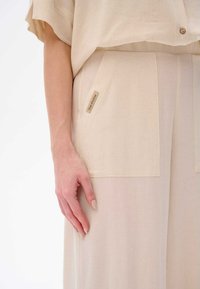 Beige Leinenoutfit mit einer lockeren Bluse und maßgeschneiderten Hosen mit vorderen Taschen. Auffälliges Etikett "blue shadow" an der Seite.