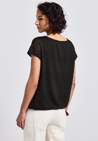 Schwarzes Mesh-Oberteil mit kurzen Ärmeln, das eine lockere Passform und ein strukturiertes Muster aufweist. Kombiniert mit hellen Hosen und damit einen lässigen Stil präsentierend.