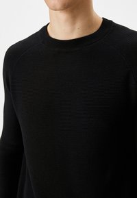 Czarny sweter o prążkowanej fakturze, okrągłym dekolcie i raglanowych rękawach. Materiał wydaje się miękki i jest prezentowany z górnej perspektywy torsu.