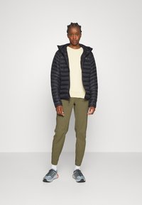 Zwarte pufferjas met capuchon, lichtbeige langetermijnshirt, groene tapered broeken en grijze sportschoenen met blauwe accenten.