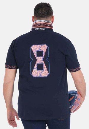 Homme portant un maillot de rugby maori bleu marine avec le numéro 8 dans le dos, tenant un ballon de rugby assorti dans la main droite, tourné de dos à la caméra.