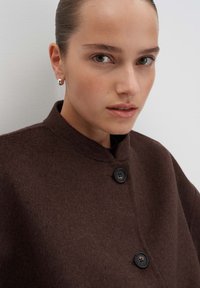 Jonge vrouw met gladgestreken haar, gekleed in een donkerbruine wolken met knopen en zilveren creolen, kijkt iets opzij.