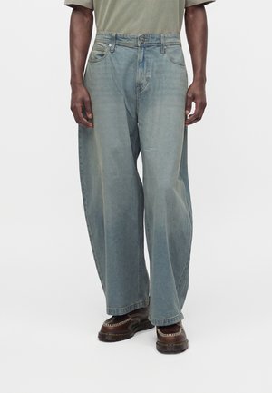 Homme portant un jean large bleu clair et des mocassins en cuir marron, debout devant un fond uni.