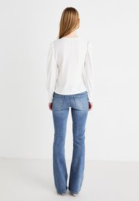 Blusa blanca texturizada con mangas abullonadas y detalles de ojal, combinada con jeans de campana de color azul claro. Los jeans tienen bolsillos traseros.
