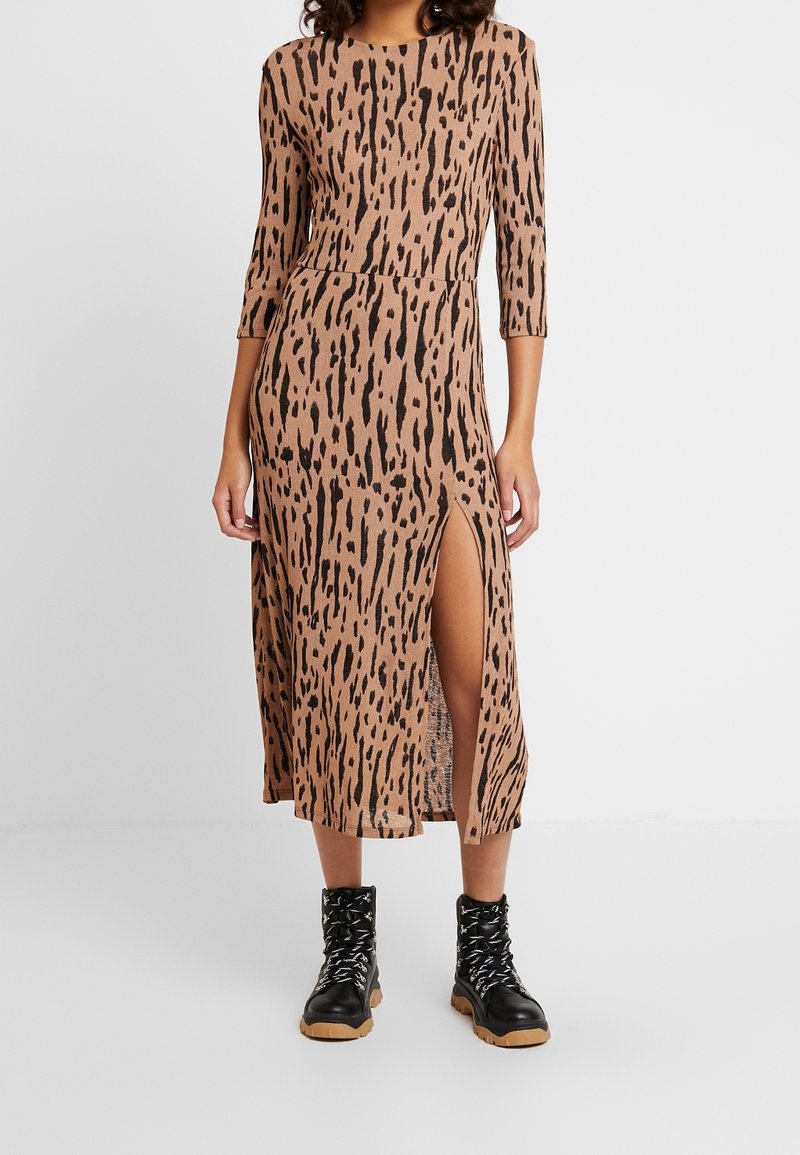 Femme portant une robe midi marron avec des taches abstraites noires, manches trois-quarts, fente latérale, et des bottes noires à lacets avec semelles couleur cuir.