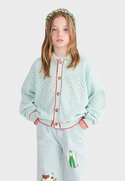 Jeune personne aux longs cheveux portant un cardigan tricoté bleu clair avec des boutons marron et un pantalon en denim clair orné d'une broderie de fermier et de vache.