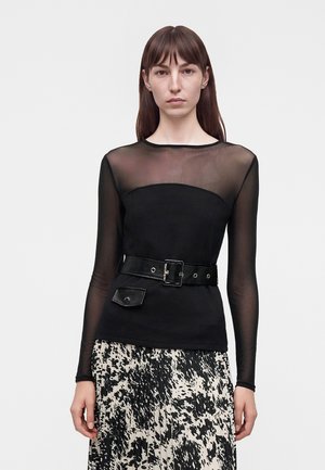 Femme portant un haut noir transparent à manches longues avec une ceinture et une jupe plissée à motifs noir et blanc, debout devant un fond uni.