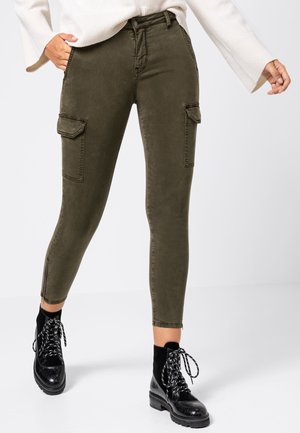 Vrouw draagt olijfgroene cargobroek met klepzakken, een wit longsleeve trui en zwarte veterlaarzen tot de enkels.