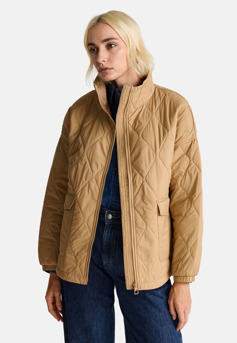 Gepolsterte braune Jacke mit Stehkragen, Reißverschluss, zwei Fronttaschen und gerippten Bündchen. Getragen über einem dunklen Jeansoutfit.