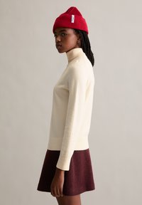 Cremfarbener Rollkragenpullover mit gerippten Manschetten, kombiniert mit einem bordeauxfarbenen, ausgestellten Rock. Das Model trägt eine rote Strickmütze mit Etikett. Schlichte, strukturierte Outfit.