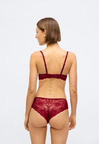 Γυναίκα που απεικονίζεται από πίσω φορώντας σκούρο κόκκινο δαντελένιο σουτιέν και ταιριαστά δαντελένια εσώρουχα σε ουδέτερο φωτεινό φόντο.