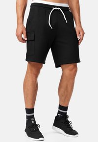 Shorts noirs en tissu doux avec une taille élastique, un cordon de serrage ajustable et une poche latérale. Portés avec des baskets noires et des chaussettes.