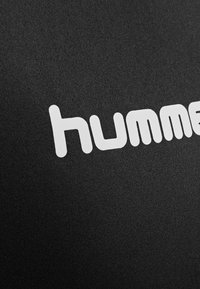 Zbliżenie białego logo "hummel" nadrukowanego na czarnej teksturowanej tkaninie.