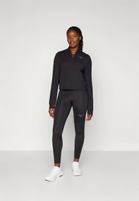 Czarny sweter z suwakiem do połowy długości i małym logo na piersi, zestawiony z czarnymi legginsami z subtelnym brandingiem, noszony z białymi trampkami.