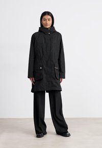 Zwarte parka-jas met capuchon, voorkant met rits, twee grote zakken en gouden accenten, gemaakt van een gladde, waterafstotende stof.