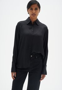Schwarze Hemdbluse mit langen Ärmeln und Kragen, aus glattem Stoff. Mit geknöpften Manschetten und entspannter Passform. Getragen mit schwarzen Jeans.