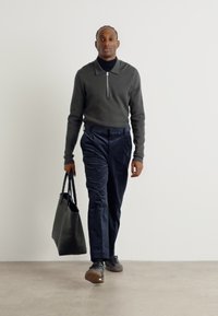 Un sweat à zip gris, un pantalon en velours côtelé bleu marine et des baskets sombres. Portant un sac fourre-tout noir. Design simple et moderne avec une coupe décontractée.