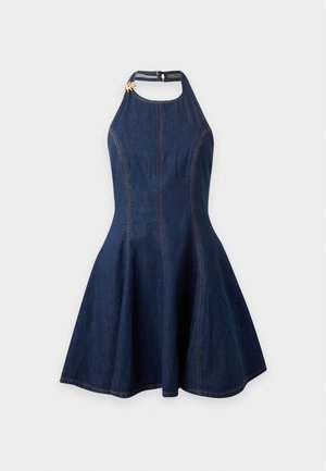 Vrouw draagt een mouwloze donkerblauwe jurk, zwarte hoge hakken en een zwarte crossbodytas met franjes, staand tegen een witte achtergrond.