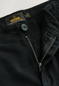 Čierne nohavice s čiastočne rozopnutým kovovým zipsom a gombíkom, so štítkom "NEXT Durable Goods" vo vnútri pásu.