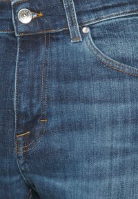 Denimjeans i mörkblått med en slät textur. Funktioner inkluderar orange sömmar, en silverknapp och subtila fickdetaljer.