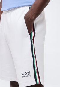 Άνδρας φοράει λευκά σορτς Emporio Armani EA7 με κόκκινη, λευκή και πράσινη ρίγα στο πλάι, χέρι στην δεξιά τσέπη και γαλάζιο πουκάμισο.