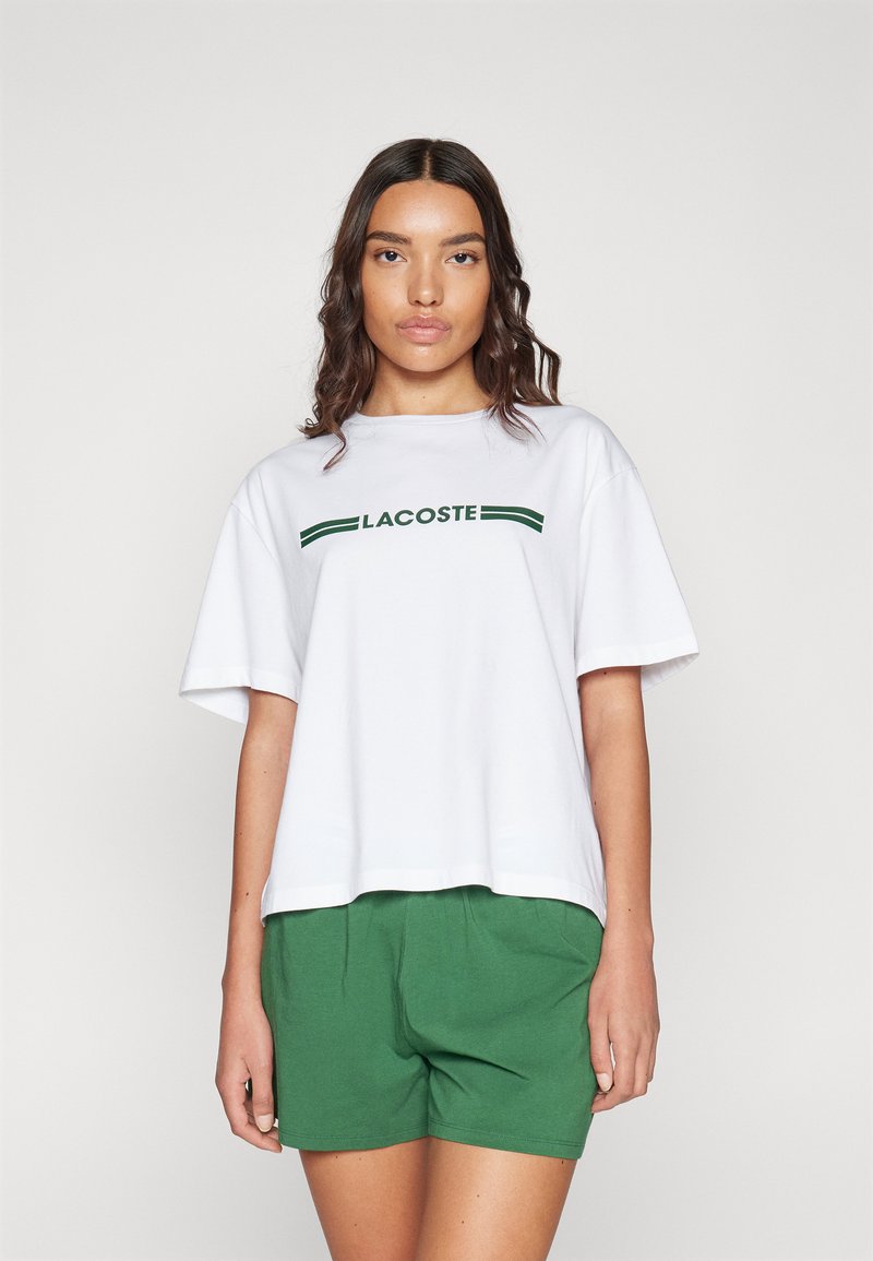 T-shirt en coton blanc à manches courtes et col rond, arborant un logo "LACOSTE" vert et deux rayures horizontales vertes en dessous. Associé à un short vert.